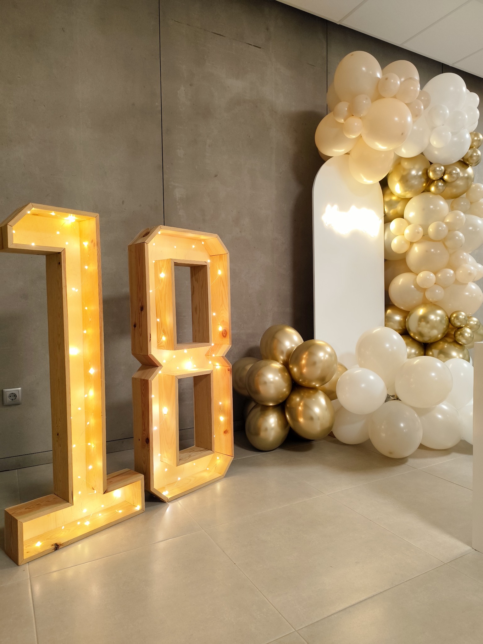 Números 18 iluminados junto a uma composição de balões brancos e dourados