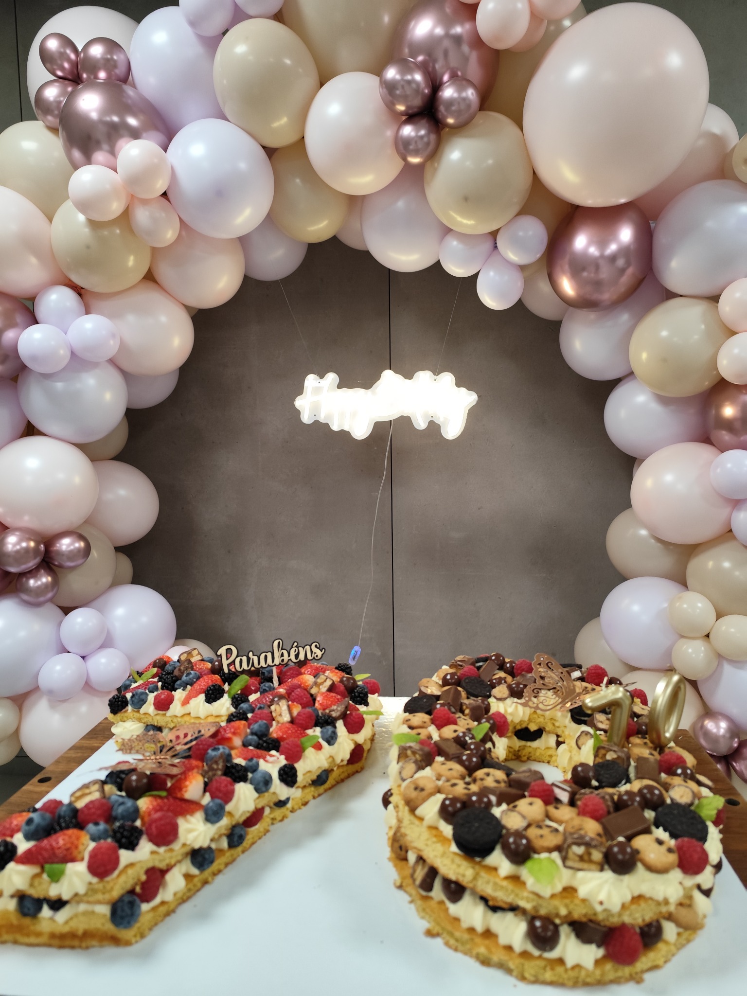 Bolos decorados com fruta e doces, diante de um arco de balões em tons suaves