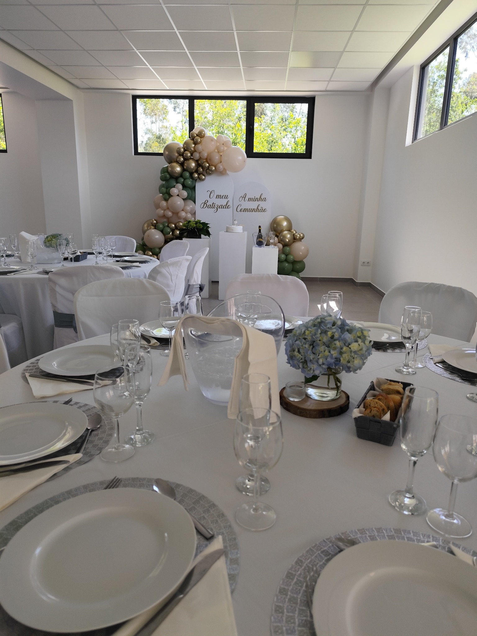 Sala preparada com mesas brancas, flores e decoração de balões para um batizado e uma comunhão