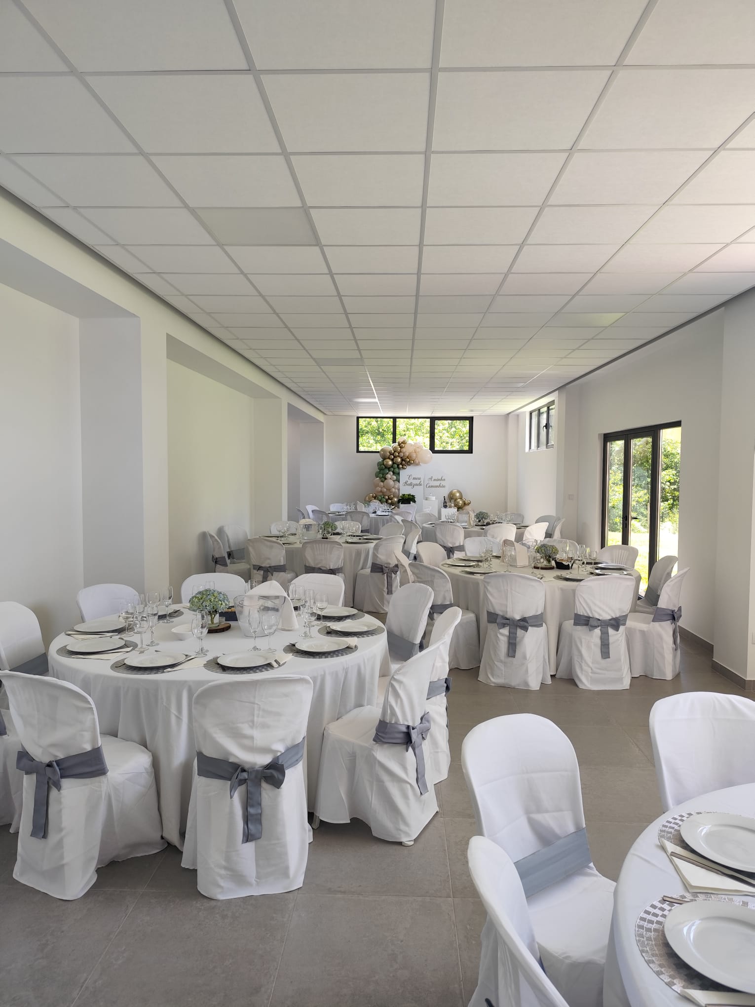 Sala do Momentos Únicos com luz natural, mesas redondas e cadeiras brancas com laços cinzentos